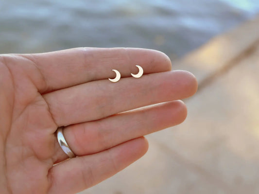 womens hand holding pair of little gold fill crescent moon stud earrings with water in background